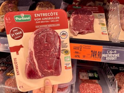 Ruka držící balený hovězí steak v supermarketu před regálem masných výrobků