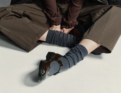 Modelka v hnědém outfitu, černých balerínách a vysokých šedých ponožkách sedí se skříženýma nohama v ateliéru
