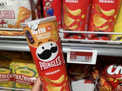 Ruka držící balení Pringles před regálem se slanými pochutinami v supermarketu