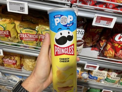 Ruka držící balení Pringles před regálem se slanými pochutinami v supermarketu