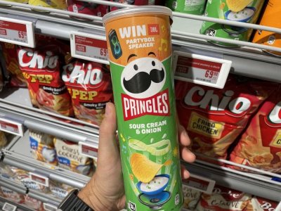 Ruka držící balení Pringles před regálem se slanými pochutinami v supermarketu