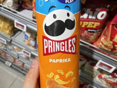 Ruka držící balení Pringles před regálem se slanými pochutinami v supermarketu