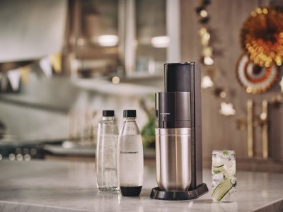Černo-stříbrný výrobník SodaStream, dvě lahve SodaStream, sklenice s vodou, ledem a okurkou
