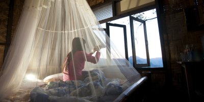 Dívka sedí v posteli přikryté moskytiérou a dívá se z okna na hory