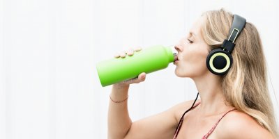 Žena se sluchátky na hlavě pije vodu ze zelené znovupoužitelné lahve