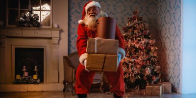 Santa Claus nese tři velké dárky položené na sobě, v pozadí krb a vánoční strom