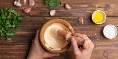 Ruce chystají majonézu v dřevěném hmoždíři, v pozadí bylinky, česnek a olej se solí