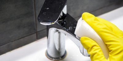 Ruka v žluté rukavici umývá bílou houbičkou vodní kohoutek.