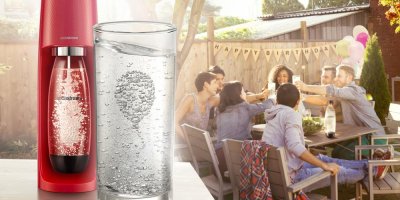 Výrobník sodovky od značky SodaStream, sklenice s perlivou vodou a v pozadí parta přátel u stolu na oslavě narozenin