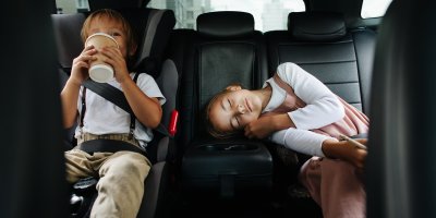 Dvě děti v autě – jedno pije z kelímku, druhé spí