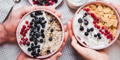 Ruce drží misku s jogurtem, borůvkami, rybízem a vločkami a druhou s jogurtem, rybízem, borůvkami a cornflakes