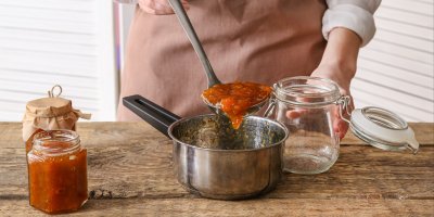 Žena přendává naběračkou broskvovou marmeládu z kastrolu do zavařovací sklenice