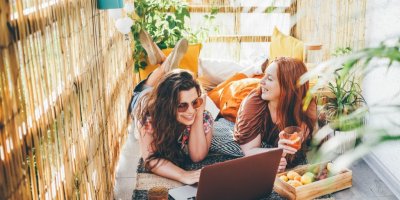 Dívky leží na balkoně s rákosovou zástěnou a koukají se na notebook