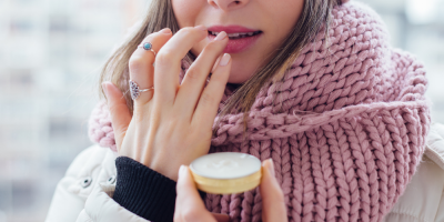 Žena v zimní bundě a šále si na rty aplikuje balzám z kalíšku