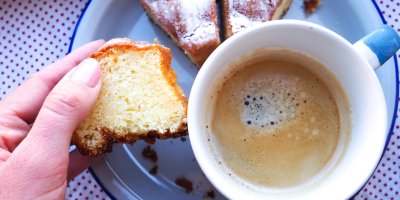 Žena drží kousek tvarohové bábovky vedle hrnečku s kávou.