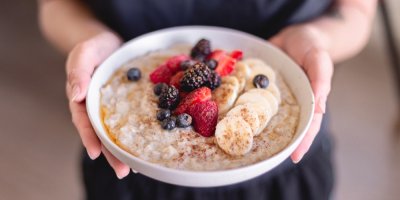 Žena drží v rukou misku s ovesnou kaší ozdobenou plátky banánu, jahod, borůvkami a ostružinami.