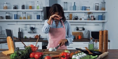 Žena si utírá oči při krájení cibule v kuchyni, kolem na stole další zelenina