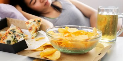 Miska chipsů, pizza a pivo, v pozadí žena ležící v posteli