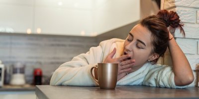 Žena v županu zívá a opírá se o stolek s hrnkem