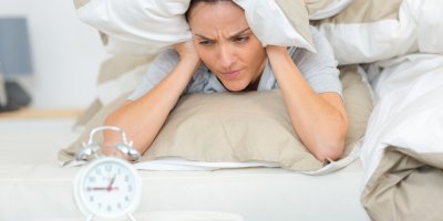 Žena v posteli s budíkem v popředí si zakrývá hlavu polštáři