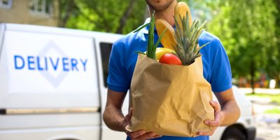 Kurýr v modrém tričku nese papírovou tašku s ovocem, zeleninou a pečivem