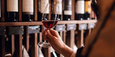 Člověk stojí ve vinárně před policí s víny, v ruce drží sklenku červeného vína