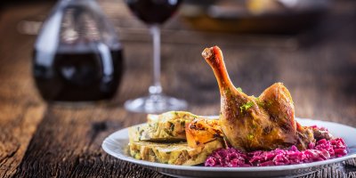 Kachní stehno, knedlíky a červené zelí na talíři, v pozadí červené víno ve džbánu a ve sklenici