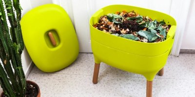Domácí kompostér limetkové barvy na čtyřech nohách naplněný hlínou a zeleninou stojí vedle kaktusu
