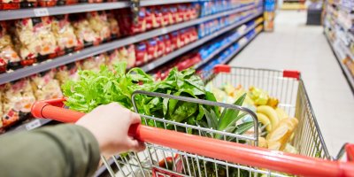 Zaostřeno na nákupní košík a dlážděnou podlahu, po stranách jsou výrobky v regálech