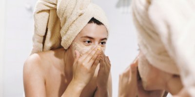 Žena má vlasy v ručníku, kouká se do zrcadla a na pleť si aplikuje peeling