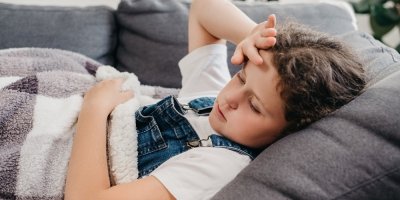 Dítě leží na gauči zabalené v dece a drží se za hlavu