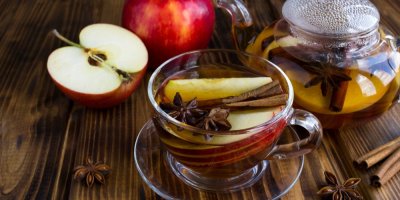 Skleněný hrnek čaje, ve kterém je celá skořice a jablko, kolem kousky jablek a skořice