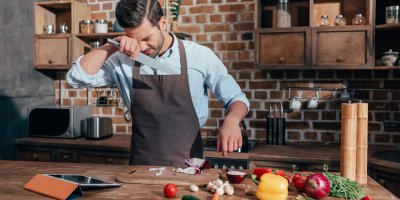 Muž v zástěře krájí červenou cibuli, kření se a přikládá si ruku k očím