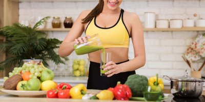 Žena si nalévá zeleninovou šťávu z mixéru do sklenice, kolem jsou papriky, brokolice, jablka, hroznové víno