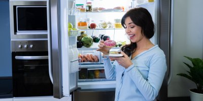 Žena stojí u otevřené lednice a v ruce drží donut, který se chystá sníst