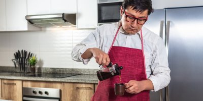 Muž v zástěře stojí v kuchyni a nalévá si kávu z french pressu do hrnku