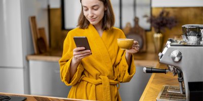 Žena v županu v jedné ruce drží mobilní telefon, ve druhé hrnek s kávou, vedle ní je pákový kávovar