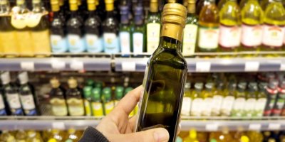 Ruka držící tmavou láhev s olejem a v pozadí regál s mnoha dalšími druhy olejů