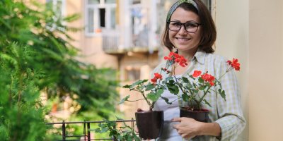 Žena stojí na balkóně a drží v ruce muškáty