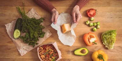 Ruce balí sýr do papíru a kolem jsou další potraviny v papíru