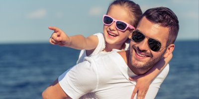 Muž má na zádech holčičku, která prstem ukazuje do dálky, oba mají sluneční brýle a za nimi je moře