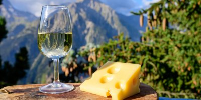 Kousek ementálu, sklenička bílého vína, v pozadí jehličnan a hory