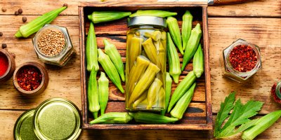 Nakládaná okra ve sklenici, kolem jsou čerstvé okry a koření