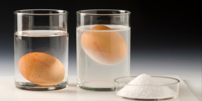 Dvě sklenice s vodou a vejci, jedno vejce je na dně, druhé se drží u povrchu, vedle hromádka soli