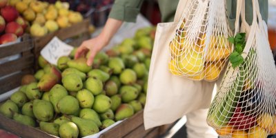 Žena drží plátěnou tašku a dvě síťovky a sahá pro hrušku
