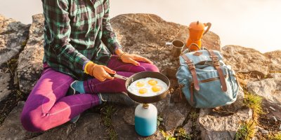 Žena sedí na hoře a připravuje si volská oka na pánvičce, vedle sebe má batoh, plechový hrnek a moka konvičku