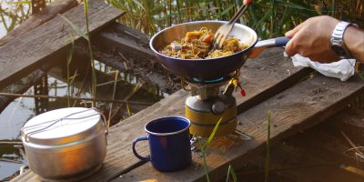 Člověk si připravuje nudle na pánvi, vedle je ešus a plechový hrnek