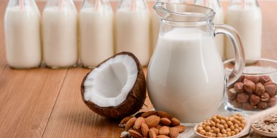 Džbán a sklenice s veganským „mlékem“ a ingredience k jeho výrobě – kokos, sója, mandle a oves