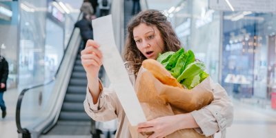 Šokovaná žena se s nákupem v ruce dívá na dlouhou účtenku