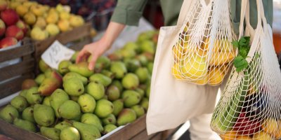 Žena si v obchodě vybírá hrušky a nese dvě síťovky plné ovoce a zeleniny